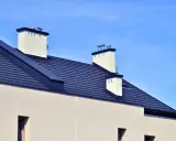 Nowoczesny dach bezokapowy z ciemnogranatową dachówką i białymi kominami, pod błękitnym niebem.