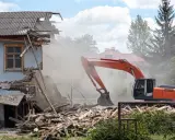 Pomarańczowy koparka prowadzi rozbiórkę budynku gospodarczego, wzbijając tumany kurzu. Ruiny i drewno leżą na ziemi.