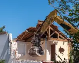 Żółta koparka z chwytakiem pracuje przy rozbiórce domu. Widać zniszczony dach i ściany.