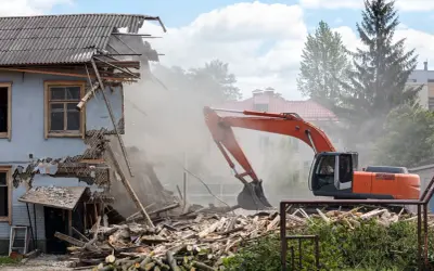 Pomarańczowy koparka prowadzi rozbiórkę budynku gospodarczego, wzbijając tumany kurzu. Ruiny i drewno leżą na ziemi.