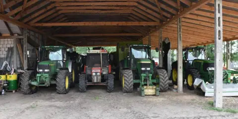 Kilka zielonych i jeden czerwony traktor stoją pod drewnianą wiatą. To pewnie ich tymczasowe schronienie, bo taka wiata bez pozwolenia to nie najlepszy pomysł.