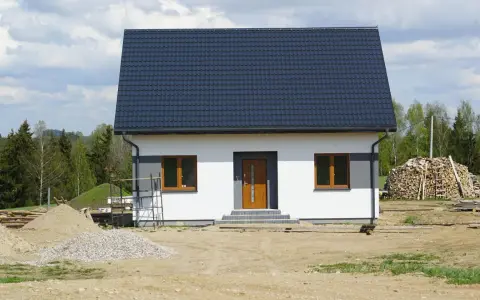 Nowy dom z ciemnym dachem i białymi ścianami, gotowy na zawiadomienie o zakończeniu budowy. Wokół plac budowy z piaskiem i drewnem.