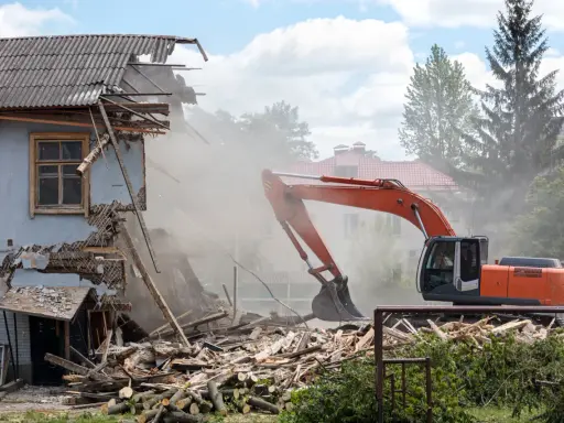 Pomarańczowy koparka prowadzi rozbiórkę budynku gospodarczego, wzbijając tumany kurzu. Ruiny i drewno leżą na ziemi.