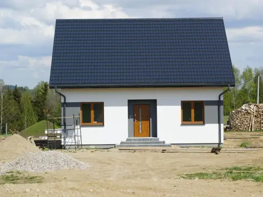 Nowy dom z ciemnym dachem i białymi ścianami, gotowy na zawiadomienie o zakończeniu budowy. Wokół plac budowy z piaskiem i drewnem.
