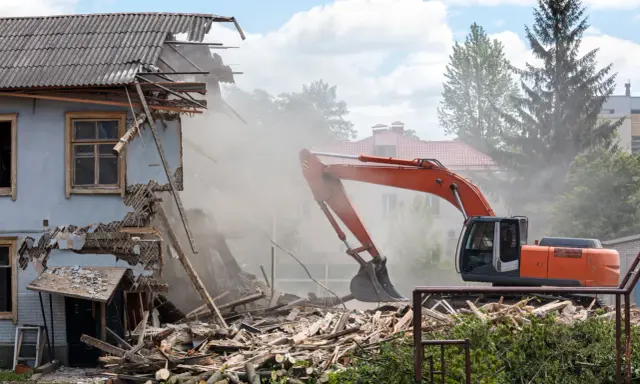 Pomarańczowy koparka prowadzi rozbiórkę budynku gospodarczego, wzbijając tumany kurzu. Ruiny i drewno leżą na ziemi.
