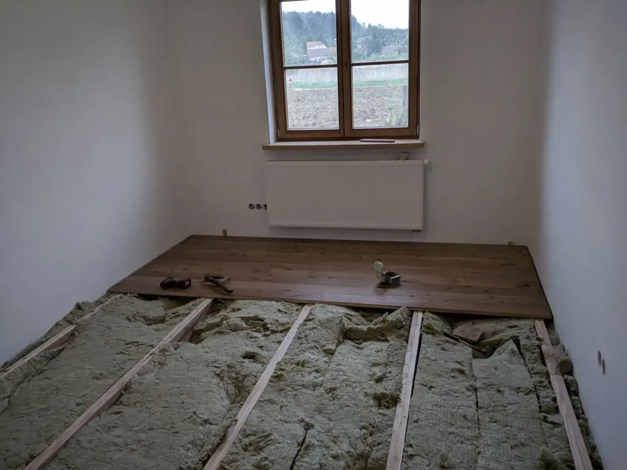 Podłoga w trakcie remontu. Widoczne drewniane legary, wełna mineralna i deski parkietowe. Narzędzia czekają na dokończenie pracy.