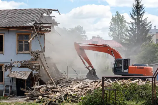 Pomarańczowy koparka prowadzi rozbiórkę budynku gospodarczego, wzbijając tumany kurzu. Ruiny i drewno leżą na ziemi.