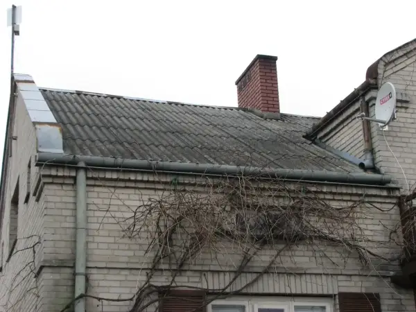 Stary dach z azbestu, pokryty falistymi płytami, z kominem i anteną satelitarną. Pnącza oplatają ścianę.