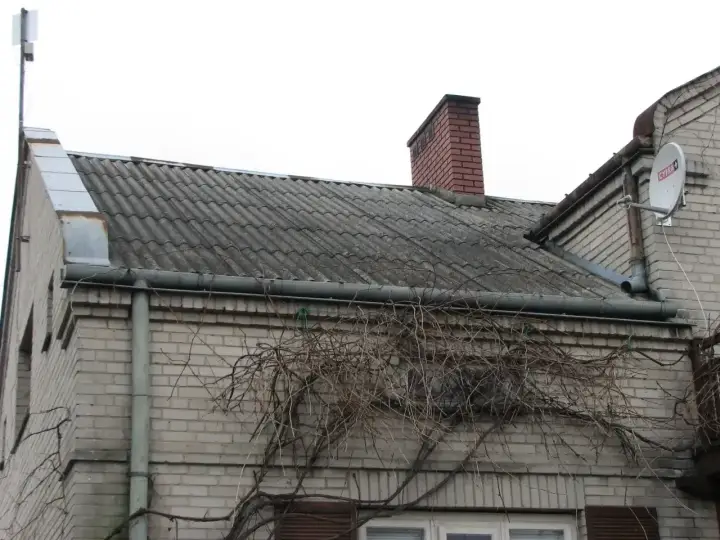 Stary dach z azbestu, pokryty falistymi płytami, z kominem i anteną satelitarną. Pnącza oplatają ścianę.