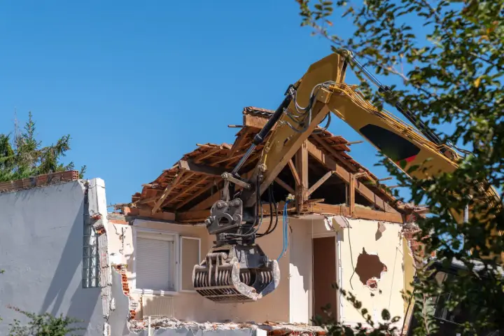 Żółta koparka z chwytakiem pracuje przy rozbiórce domu. Widać zniszczony dach i ściany.
